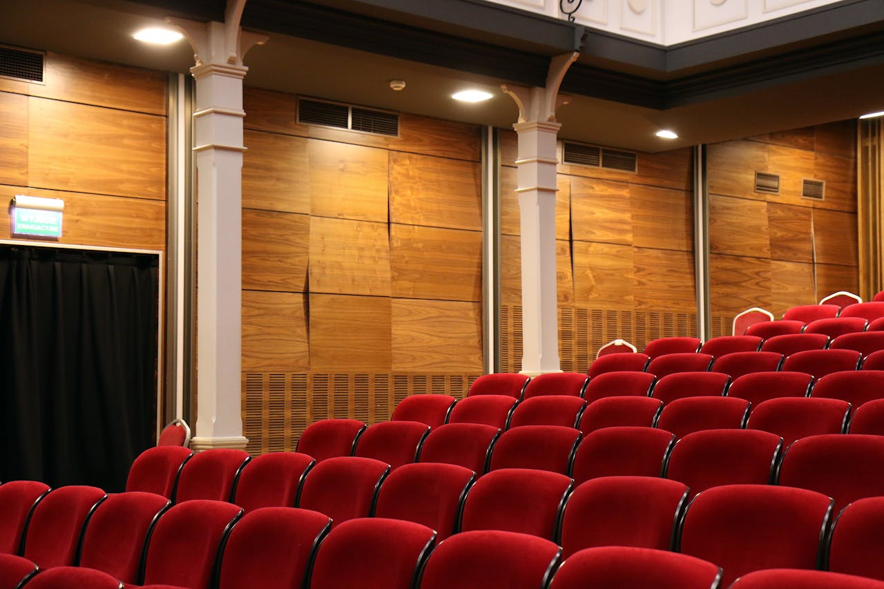 home-hero Interior of an empty auditorium with red seats and wooden panels, lit overhead.
