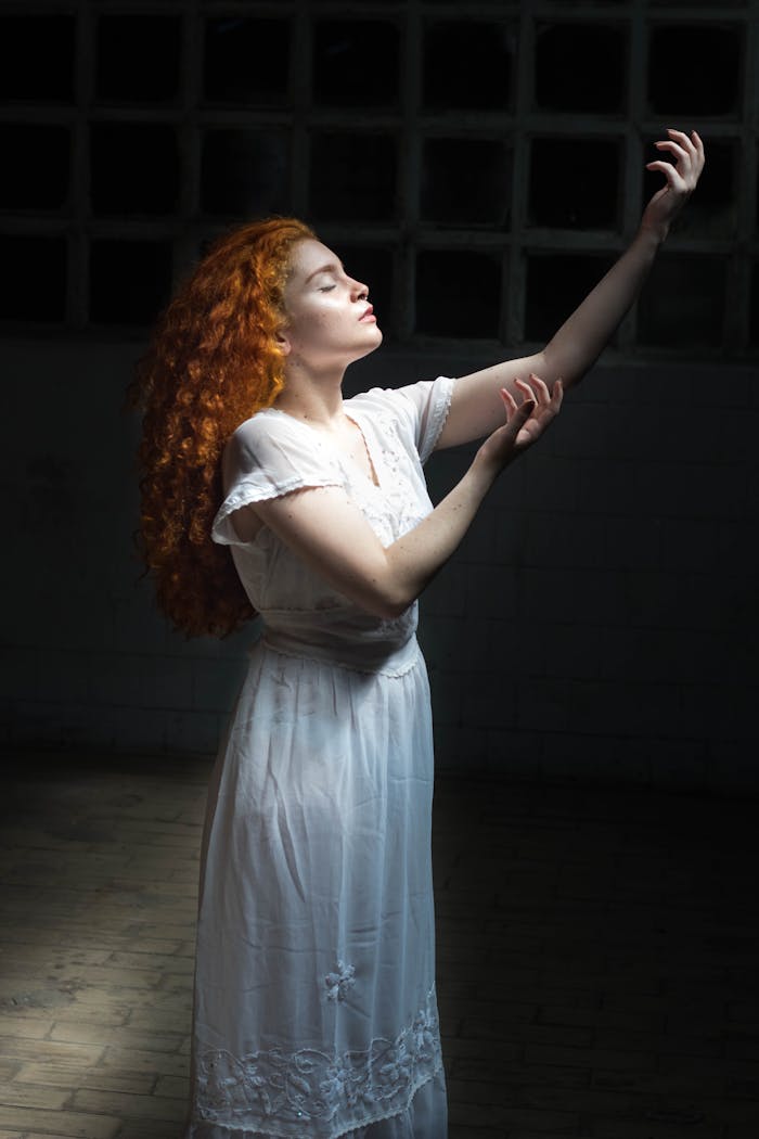why-choose-us A young red-haired woman in a white dress posed gracefully with dramatic lighting creating an artistic portrait.
